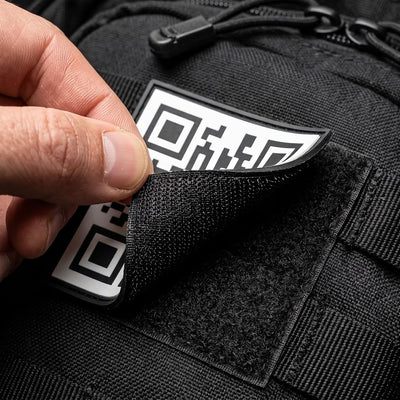 A close-up of a hand peeling back a black textured patch with a Rick Roll QR code from a black tactical bag or backpack.